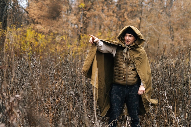 Retrato de turista masculino vestindo barraca de capa de chuva verde em pé no matagal de arbustos em dia nublado frio e apontando o dedo na direção. Conceito de pesquisa, viagem e sobrevivência.