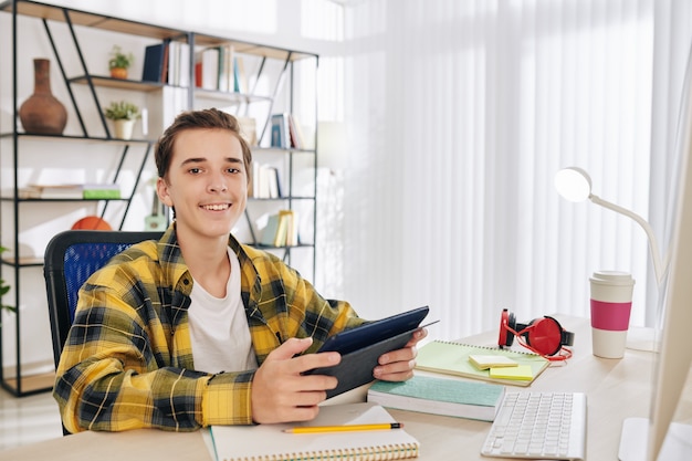 Retrato de um adolescente alegre sentado em sua mesa com um tablet digital nas mãos e sorrindo para a câmera