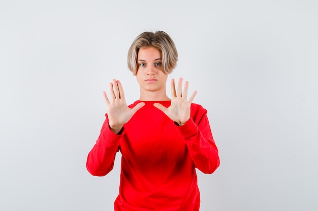 Retrato de um adolescente loiro mostrando um gesto de parada no suéter vermelho e olhando confiante para a frente