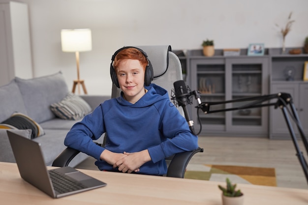 Retrato de um adolescente ruivo usando fones de ouvido e sorrindo para a câmera enquanto está sentado na mesa com o microfone configurado para podcasting ou streaming online, copie o espaço