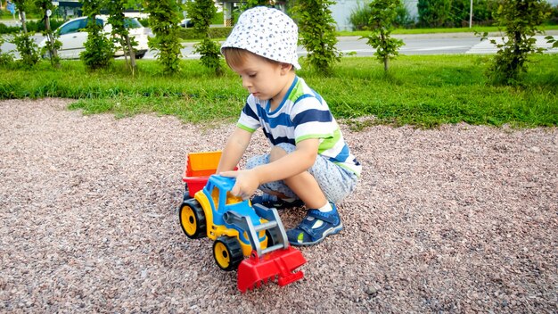 Retrato de um adorável menino de 3 anos brincando com um caminhão de brinquedo com reboque no parquinho do parque