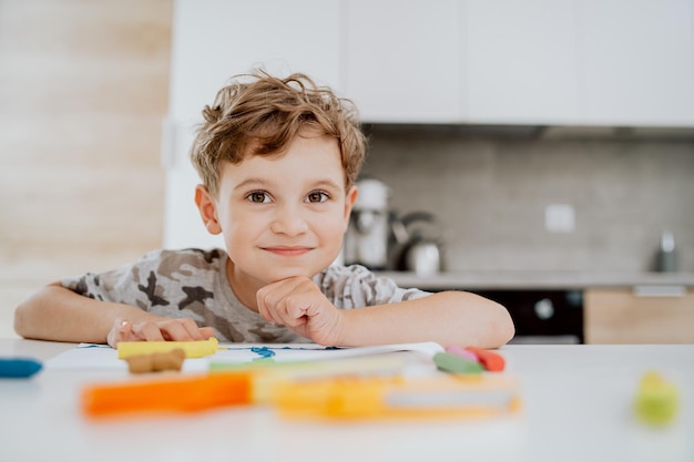 Retrato de um adorável menino escolar feliz sentado à mesa da cozinha aproveitando algum tempo livre pouco