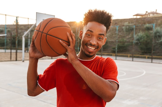 Retrato de um africano feliz segurando uma bola enquanto joga basquete no parquinho ao ar livre