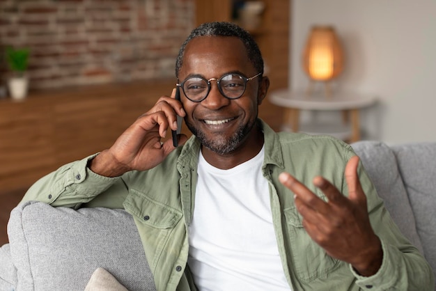 Retrato de um alegre homem afro-americano de meia-idade em chamadas de óculos por smartphone gesticula fala