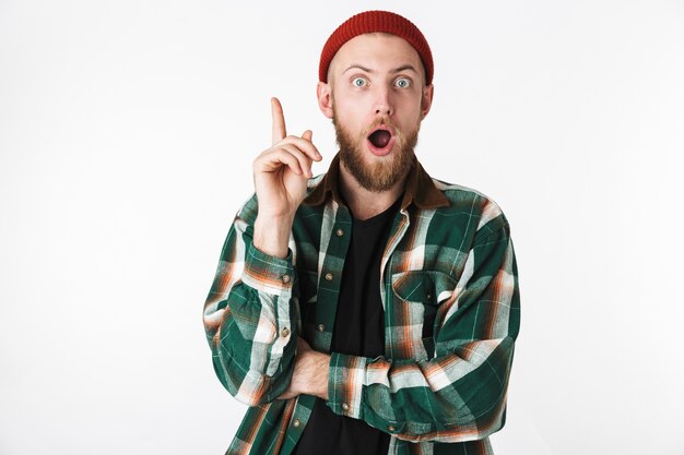 Retrato de um barbudo surpreso com chapéu e camisa xadrez apontando o dedo para cima, em pé, isolado sobre um fundo branco