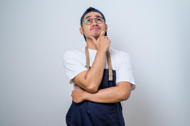 Retrato de um barista asiático atraente em avental azul pensando em questão com a mão no queixo