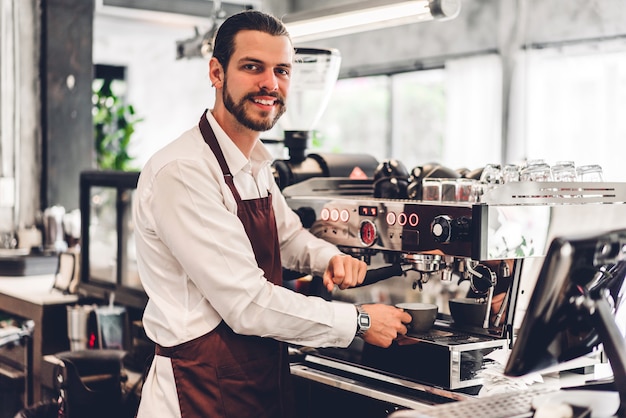 Retrato de um barista barbudo bonito proprietário de uma pequena empresa trabalhando usando a máquina de café para fazer café atrás do balcão de um café