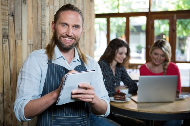 Retrato de um barista masculino feliz escrevendo ordens para clientes do sexo feminino em segundo plano em uma cafeteria