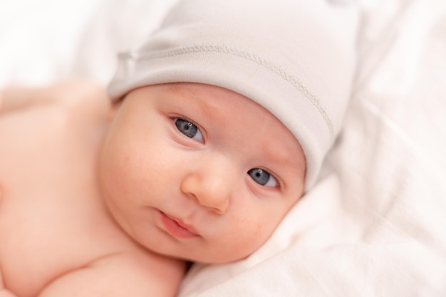 Retrato de um bebê com olhos azuis em uma fralda e um boné branco deitado de costas na roupa de cama branca. espaço para texto. Foto de alta qualidade