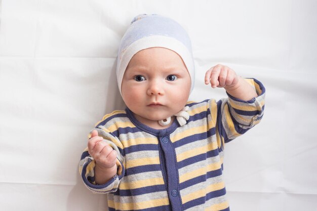 Retrato de um bebê com roupas com uma cara de expressão engraçada no lençol branco. Camada plana, vista superior.