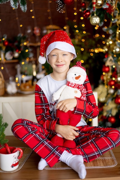 Retrato de um bebê feliz sentado em uma mesa de cozinha com uma decoração de Natal vermelha com um brinquedo de boneco de neve e sorrindo em antecipação ao feriado