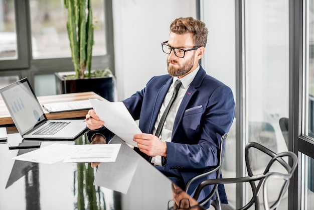 Retrato de um belo banqueiro trabalhando com um laptop sentado no interior do escritório de luxo com uma bela vista sobre os arranha-céus