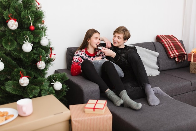 Retrato de um belo casal sentado no sofá em casa e comendo chocolate enquanto passa tempo juntos perto da árvore de Natal