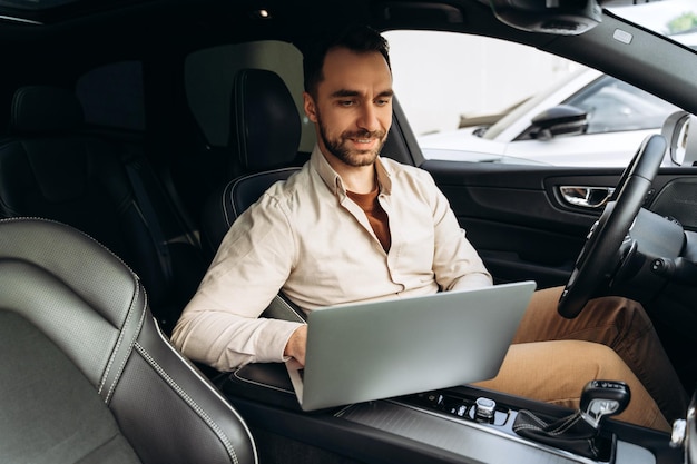 Retrato de um belo empresário feliz, um empregado profissional sentado em um carro novo usando um laptop