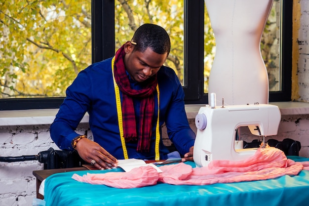 Retrato de um belo homem africano costureira sorridente com máquina de costura. Designer elegante de homem afro-americano trabalhando em um manequim de oficina de alfaiate, fita métrica de mesa na sala contra a janela de outono