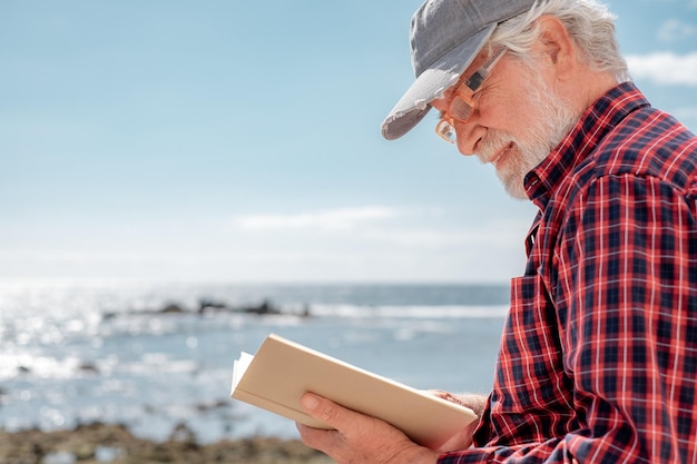 Retrato de um belo homem barbudo sênior lendo um livro em frente ao mar aproveitando a aposentadoria