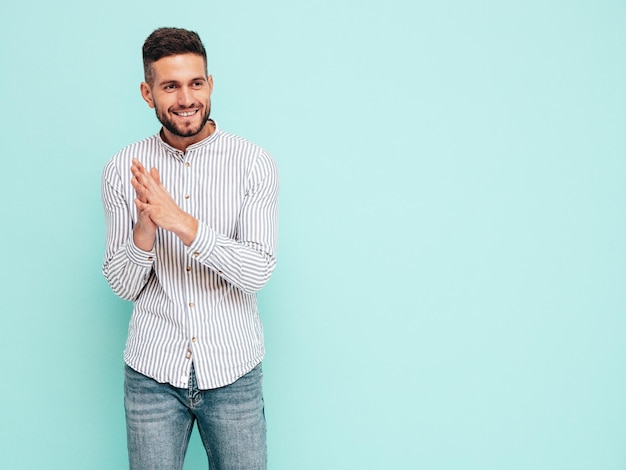 Retrato de um belo modelo sorridente Sexy homem elegante vestido de camisa e jeans Moda hipster masculino posando perto da parede azul no estúdio Alegre e feliz