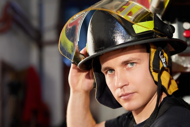 Retrato de um bombeiro bonito musculoso em um fundo de caminhão de bombeiros segurando um capacete de bombeiros olhando para o lado