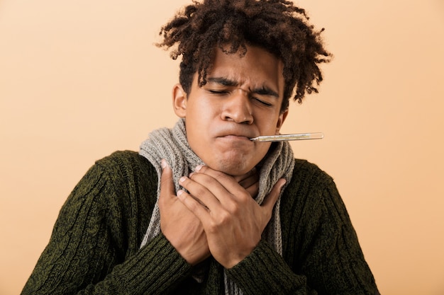 Foto retrato de um cara afro-americano doente, vestindo um suéter e um lenço, agarrando a garganta com um termômetro na boca, isolado sobre uma parede bege