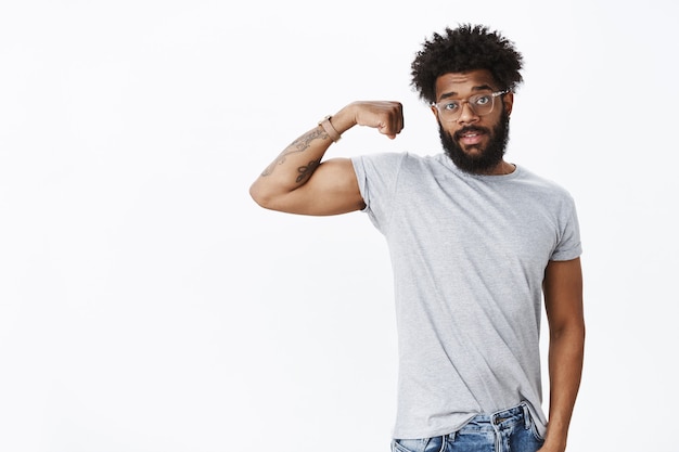 Retrato de um cara barbudo comum afro-americano bonito com cabelo encaracolado em óculos, levantando uma mão e exibindo bíceps sendo fortes enquanto se exercita na academia convidando as pessoas a entrarem