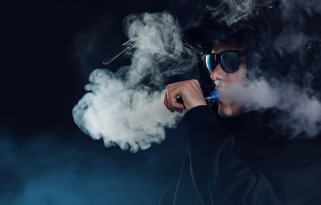 Retrato de um cara bonito com um chapéu preto e óculos escuros, vaporizando e exalando uma nuvem de vapor de um cigarro eletrônico