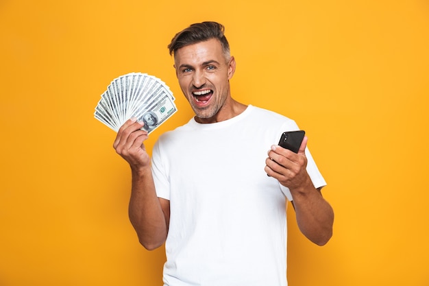 Retrato de um cara de 30 anos com a barba por fazer, uma camiseta branca segurando um celular e um monte de dinheiro isolado em amarelo