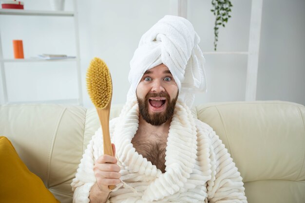 Retrato de um cara engraçado usando uma toalha de turbante segurando uma escova de massagem com as mãos enquanto está sentado na pele masculina do sofá