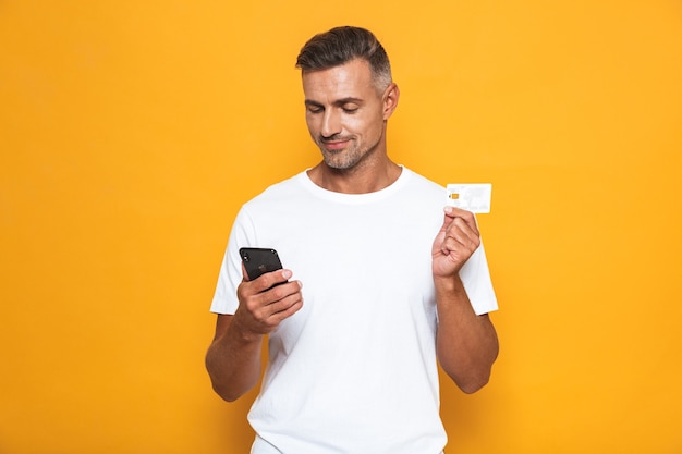 Retrato de um cara europeu dos anos 30 em uma camiseta branca segurando um celular e um cartão de crédito isolado em amarelo