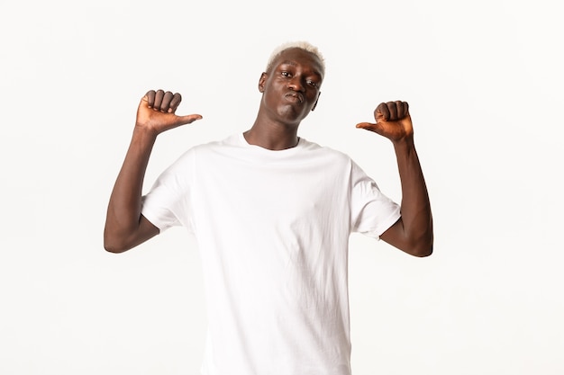 Retrato de um cara loiro afro-americano bonito e confiante, apontando o dedo para si mesmo atrevido e orgulhoso, em pé sobre um fundo branco