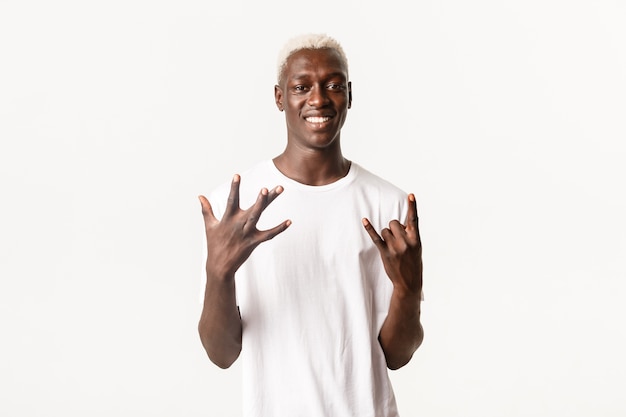 Retrato de um cara loiro afro-americano elegante mostrando sinal de mão da costa oeste e rock-on, sorrindo otimista