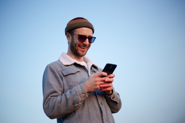 Retrato de um cara sorridente moderno com o smartphone nas mãos no fundo do céu azul