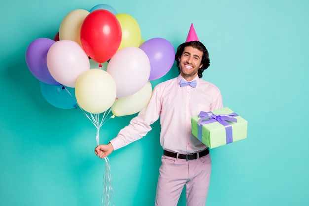 Retrato de um cara sorridente segurando uma caixa de presente de balões coloridos vestida de casquinha formal isolada em um fundo de cor turquesa