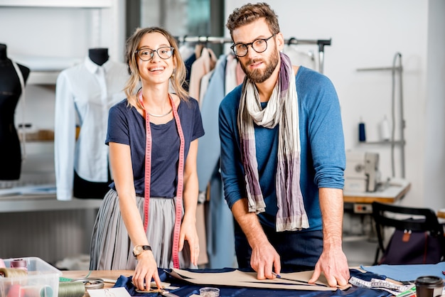 Retrato de um casal de designers de moda trabalhando com esboços de tecidos e roupas em um estúdio cheio de ferramentas e equipamentos de alfaiataria