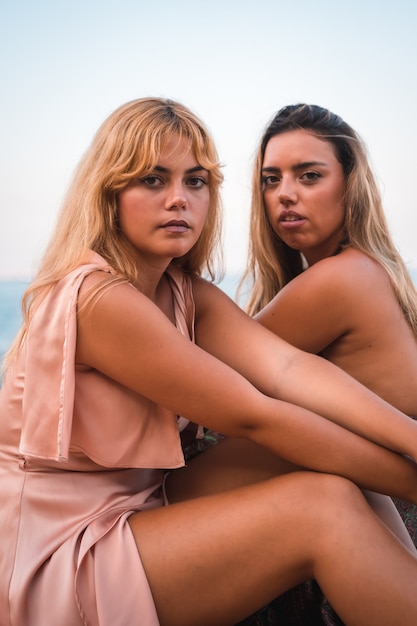 Foto retrato de um casal de garotas caucasianas se divertindo sentadas em vestidos muito bonitos, aproveitando o verão à beira-mar