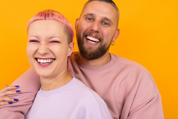 Retrato de um casal feliz de marido e mulher com cabelos coloridos e piercings vestidos com roupas cor-de-rosa caasual está de pé amarelo
