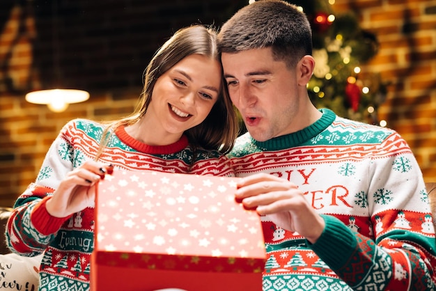 Retrato de um casal romântico abrindo uma caixa de presente à noite perto da árvore de natal decorada