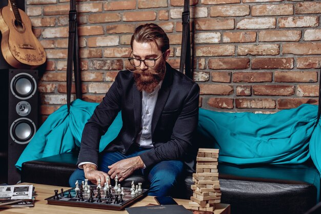 Retrato de um cavalheiro inteligente inteligente com óculos, bigode e barba, sentado no sofá e jogando xadrez.