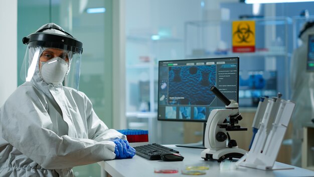 Retrato de um cientista cansado no macacão, olhando para a câmera sentada no moderno laboratório equipado. Equipe de médicos examinando a evolução do vírus usando ferramentas de alta tecnologia e química para o desenvolvimento de vacinas.