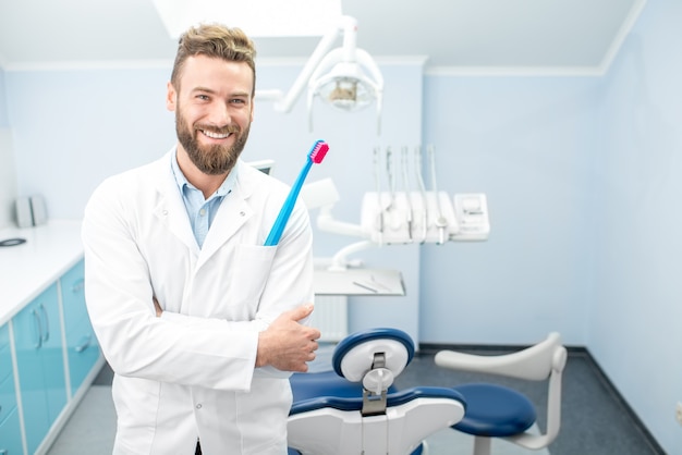 Retrato de um dentista bonito de uniforme com uma grande escova de dentes no consultório odontológico