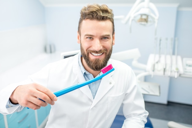 Retrato de um dentista engraçado de uniforme com uma grande escova de dentes no consultório odontológico