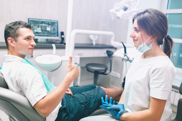 Retrato de um dentista fêmea e de um paciente masculino feliz novo em um consultório dentista. homem desistindo polegares