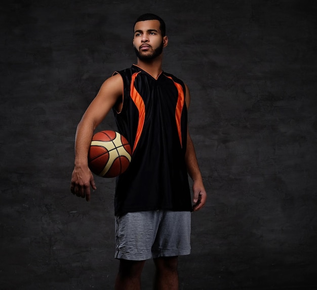 Retrato de um desportista afro-americano. Jogador de basquete em roupas esportivas com uma bola em um fundo escuro.