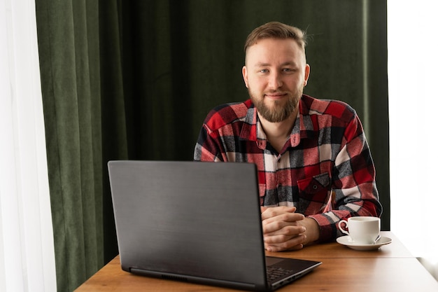 Retrato de um empregado feliz sorrindo para a câmera enquanto trabalha em um laptop em um escritório doméstico ou café