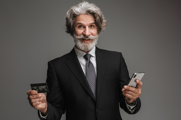 Retrato de um empresário adulto animado, vestindo um terno preto formal, segurando um cartão de crédito e um smartphone isolados sobre uma parede cinza