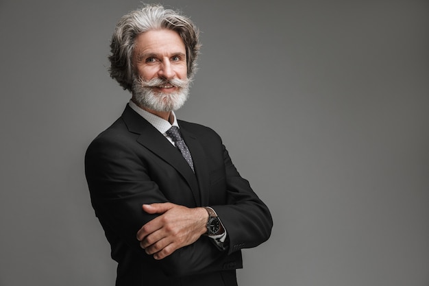 Retrato de um empresário adulto feliz vestindo um terno preto formal, sorrindo para a câmera com os braços cruzados isolados sobre a parede cinza