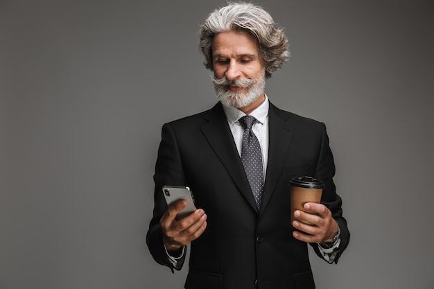 Retrato de um empresário adulto sorridente, vestindo um terno preto formal, segurando um copo de papel e um smartphone isolados sobre uma parede cinza