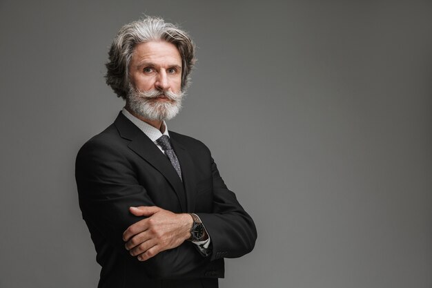 Retrato de um empresário adulto vestindo terno preto formal, olhando para a frente com os braços cruzados isolados sobre a parede cinza