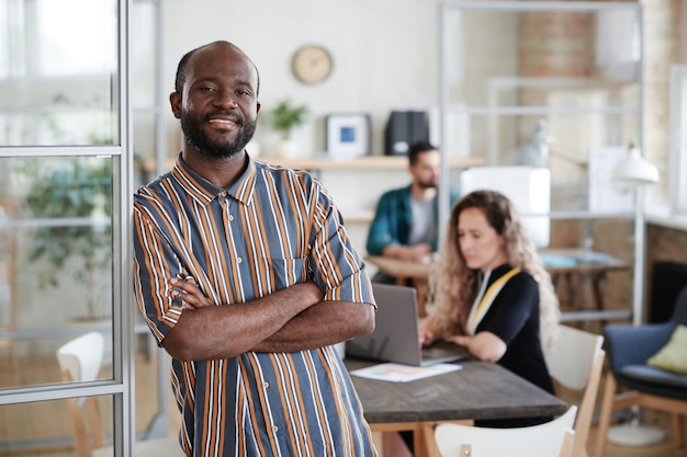 Retrato de um empresário africano barbudo em pé com os braços cruzados e sorrindo para a câmera com seus colegas trabalhando no escritório em segundo plano