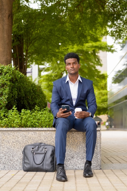 Retrato de um empresário africano usando o telefone e segurando uma xícara de café para levar ao ar livre na cidade enquanto usa o telefone celular