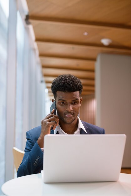 Retrato de um empresário africano usando um laptop e falando ao telefone na cafeteria, foto vertical